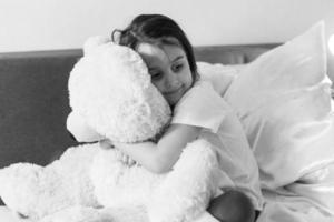 Sweet little girl is hugging a teddy bear looking at camera and smiling while sitting on her bed photo