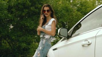 glamorös ung kvinnor i jeans klänning talande på de telefon nära henne bil på de gata video