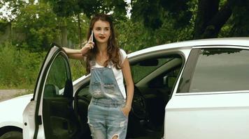 young  girl in jeans dress and glasses stands near the machine and speaks on a mobile telephone video