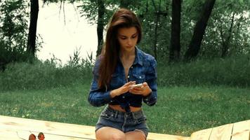 morena esbelta atraente na camisa, sentado em um banco e olhando no telefone video