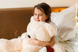 dulce niña está abrazando a un oso de peluche mirando a la cámara y sonriendo mientras está sentada en su cama foto