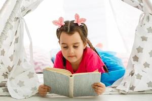 Enjoying great story. Cute little girl reading book while sitting on the floor in tent photo