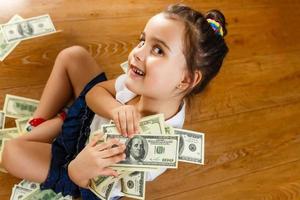 una hermosa niña juega con billetes de cien dólares, los despliega frente a ella en el suelo en un montón, foto