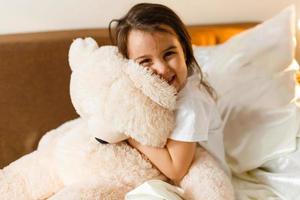 dulce niña está abrazando a un oso de peluche mirando a la cámara y sonriendo mientras está sentada en su cama 396815581 foto