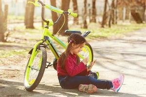 Child traveling bicycle in summer park. Bicyclist little girl watch on mobile phone. Kid counts pulse after sport training and is looking for way to navigator. photo