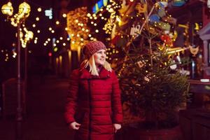 Outdoor photo of young beautiful happy smiling girl posing in street. Festive Christmas fair on background. Model wearing stylish winter coat, knitted beanie hat, scarf.