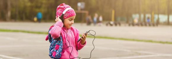 Little girl in a skirt sitting on a park bench listening to music and looking at mobile phone photo