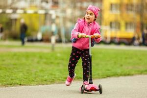 Little child learning to ride a scooter in a city park on sunny summer day. Cute preschooler girl riding a roller. Kids play outdoors. Active leisure and outdoor sport for children. photo