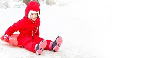 una niña muy sonriente con su traje de esquí deslizándose por una pequeña colina cubierta de nieve con su trineo foto