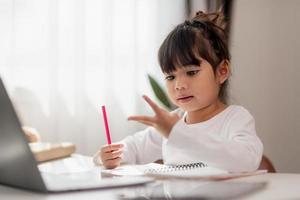 Asian schoolgirl doing her homework with laptop at home. Children use gadgets to study. Education and distance learning for kids. Homeschooling during quarantine. Stay at home photo