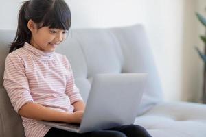 la niña asiática de la escuela usa una computadora portátil sentada en el sofá sola en casa. niño aprendiendo a leer contenido de medios sociales en línea, jugar lecciones de educación juego chatear con amigos. foto