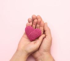 female and male hand holding a small red knitted heart on a pink background photo