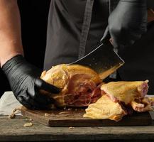 chef in black uniform and latex gloves chopping raw chicken into pieces on a brown wooden board photo