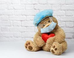 brown teddy bear sits in protective plastic glasses, a medical disposable mask and a blue cap