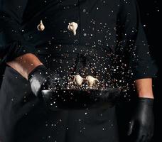 man in a black uniform holding a round cast iron pan with salt and pepper, chef tosses spice up photo