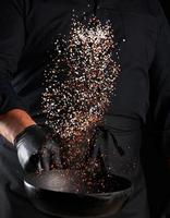 man in a black uniform holding a round cast iron pan with salt and pepper, chef tosses spice up photo