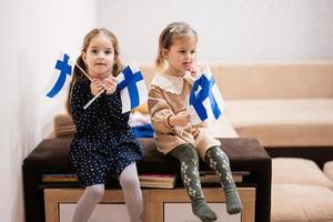 Two sisters are sitting on a couch at home with finnish flags on hands. Finland children girls with flag . photo