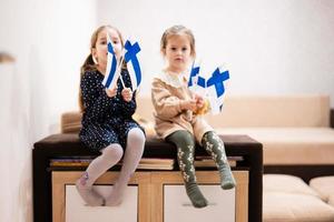 Two sisters are sitting on a couch at home with finnish flags on hands. Finland children girls with flag . photo