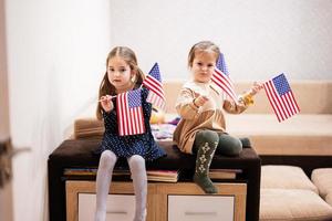 Two sisters are sitting on a couch at home with american flags on hands. USA children girls with flag . photo