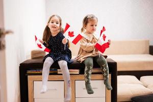 dos hermanas están sentadas en un sofá en casa con banderas canadienses en las manos. Canadá niños niñas con bandera. foto