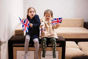 Two sisters are sitting on a couch at home with british flags on hands. Great Britain children girls with flag . photo