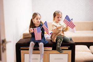 Two sisters are sitting on a couch at home with american flags on hands. USA children girls with flag . photo