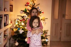 Two sisters together near Christmas tree at evening home. photo