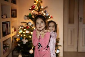 Two sisters together near Christmas tree at evening home. photo
