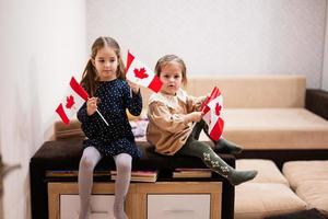 Two sisters are sitting on a couch at home with canadian flags on hands. Canada children girls with flag . photo