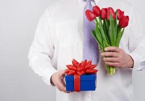 adult man in a white shirt and a lilac tie holding a bouquet of red tulips with green leaves and gift box photo