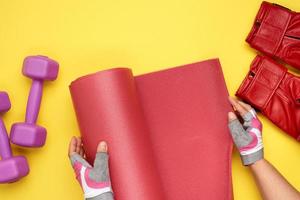 Female hands in sports gloves unfold a red sports mat on a yellow background, photo