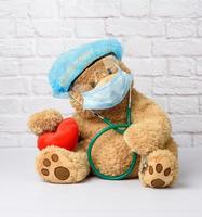 teddy bear sits in protective plastic glasses, a medical disposable mask and a blue cap photo