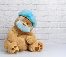 brown teddy bear sits in protective plastic glasses, a medical disposable mask and a blue cap photo