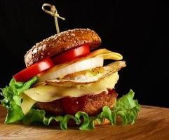 burger with meat cutlet, cheese, fried egg, tomatoes, cucumber slices and green lettuce, fast food on a round wooden board photo