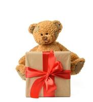 lindo oso de peluche marrón junto a una caja con un regalo atado con una cinta de seda roja foto