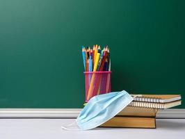 disposable medical mask, stack of books and school stationery on green chalk board background photo