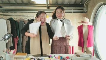 asiatico di mezza età femmina moda progettista e giovane adolescente apprendista sarto celebrare e allegro con vestito design opera successo, lanciare lavoro carta nel studio, contento costume boutique piccolo attività commerciale. video