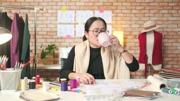 une créatrice de mode asiatique d'âge moyen travaille dans un studio de création, dessinant, buvant du café, pensant à des idées, imaginant une collection de conception de vêtements et une petite entreprise de tailleur professionnel. video
