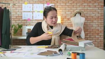 Aziatisch middelbare leeftijd vrouw mode ontwerper werken in studio, snijdend en kiezen kleding stof patroon ideeën met tekening schetsen voor jurk ontwerp collecties. professioneel winkel kleermaker mkb ondernemer. video