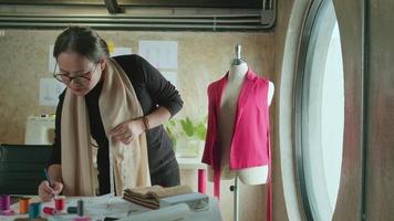 Asian middle-aged female fashion designer works in a studio by measuring puppet shapes and drawing sketches ideas, imagination dress design collection, professional boutique tailor SME entrepreneur. video