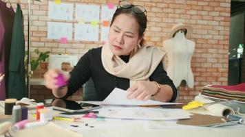 Aziatisch middelbare leeftijd vrouw mode ontwerper werken in studio door kiezen draad kleur verzameling ideeën en stijl concepten voor jurk ontwerp naaien banen. professioneel winkel kleermaker mkb ondernemer. video