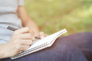 close up hand young man are using pen writing Record Lecture note pad into the book sitting on the chair outdoor. Blank book page copy  write a message. photo
