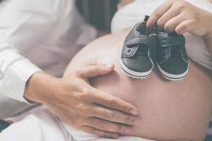 la mujer embarazada y su marido empalman la barriga sosteniendo pequeños zapatos para el bebé nonato en el vientre relajándose en casa en el dormitorio. espere la fecha de nacimiento esperando un puesto de bebé al aire libre. foto