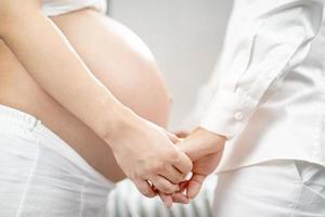 la mujer embarazada y su esposo empalman la mano abrazando la barriga en forma de corazón muestran amor por el bebé por nacer en el vientre relajándose en casa en el dormitorio. esperar la fecha de nacimiento esperando un puesto de bebé al aire libre foto