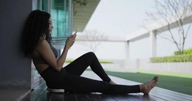 Handaufnahme, Seitenansicht einer Frau mit lockigem Haar in Sportkleidung, die auf dem Boden sitzt und das Smartphone in der Hand zum Entspannen nach dem Training im Freien des Fitnesscenters verwendet