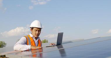 Young Asian technician man checking operation of sun and cleanliness of photovoltaic solar panel and typing on laptop computer while working in solar farm video