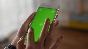 Closeup of hand woman using smartphone with green screen while sitting in living room. Blank digital smartphone in hand girl. Showing content videos blogs tapping on center screen.