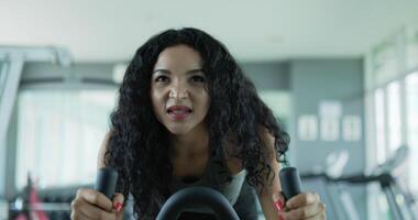 toma de mano en cámara lenta, vista frontal asiática joven máquina de bicicleta giratoria para hacer ejercicio, hermosa mujer de cabello rizado ejercicio con giro en simulador de bicicleta de aire para la salud de cardiología en el gimnasio. video