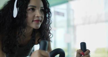 prise de vue au ralenti à la main, jeune femme asiatique dans un casque sans fil tout en faisant tourner la machine à vélo dans la salle de gym, exercice de femme aux cheveux bouclés avec spin sur simulateur de vélo à air pour la santé de la cardiologie video