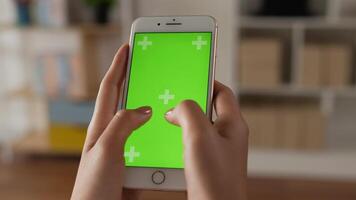 Closeup of hand woman typing smartphone with green screen while sitting in living room. Blank digital smartphone in hand girl. Showing content videos blogs tapping on center screen.